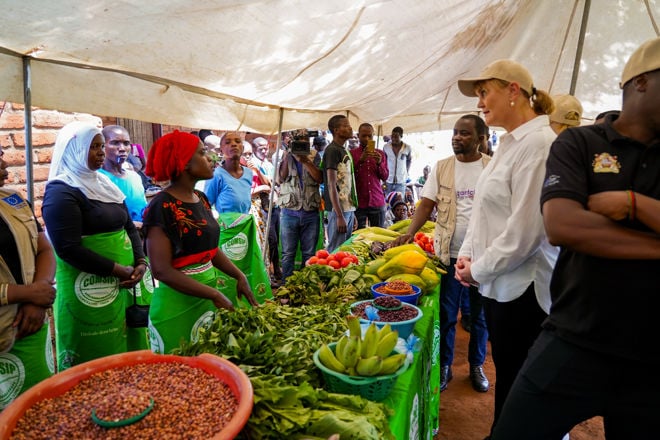 Flere kvinner og menn står samlet under et telt ved et bord med frukt og grønnsaker på et marked.