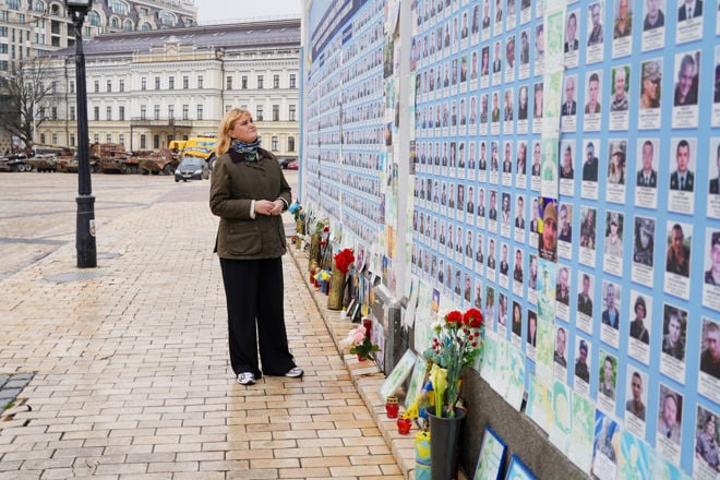En voksen kvinne med lyst hår står foran en minnevegg med bilder av mange personer og blomster plassert ved foten av veggen.