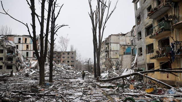 En ødelagt gate med sammenraste bygninger, trær uten blader og snø på bakken; én person går alene blant ruinene.