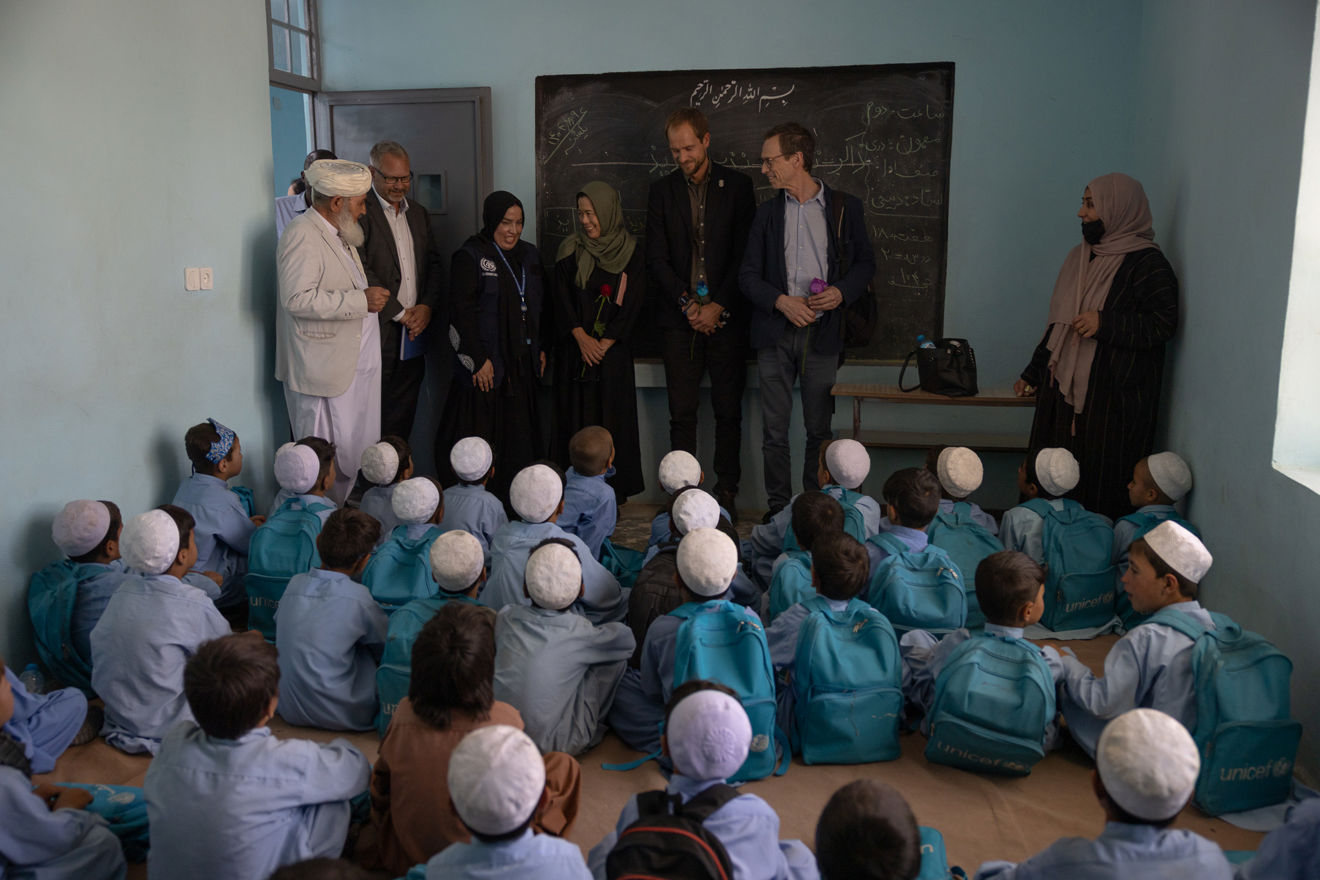 Bildet viser et klasserom med lyseblå vegger. På gulvet sitter mange barn tett i tett, alle har på seg lyseblå skoleuniformer og hvite kalotter på hodet, og de har turkise ryggsekker med UNICEF-logoen på ryggen. Barna sitter vendt mot en gruppe voksne som står foran en tavle. Blant de voksne er det både menn og kvinner, noen i tradisjonelle klær og andre i mer vestlig klesstil. 