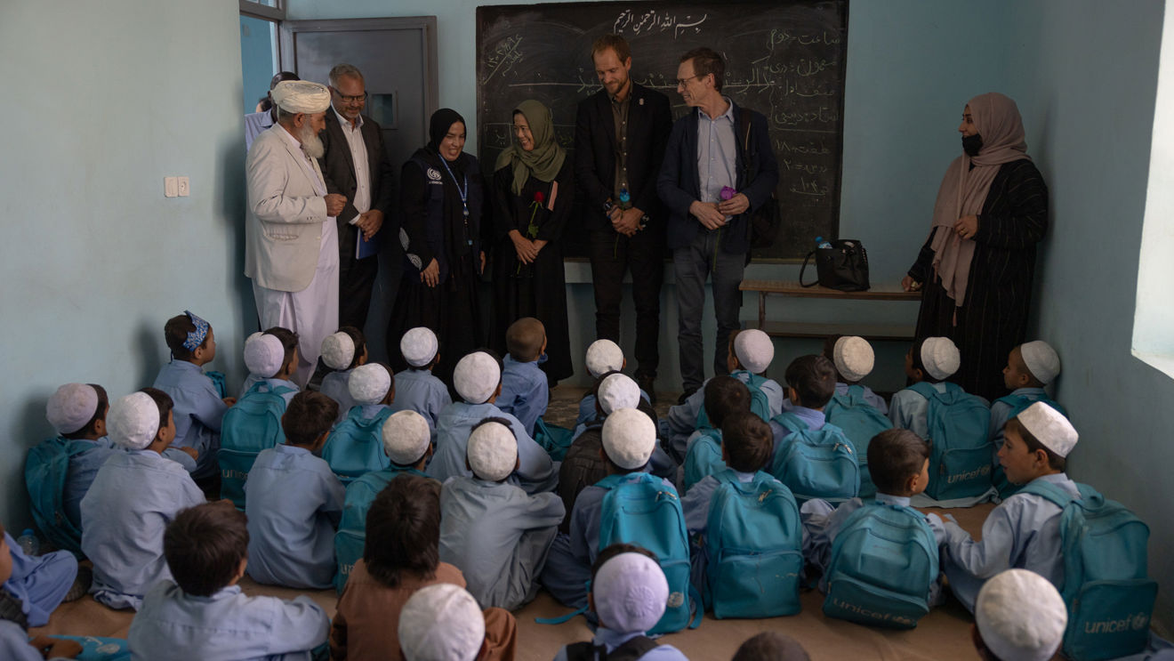 Bildet viser et klasserom med lyseblå vegger. På gulvet sitter mange barn tett i tett, alle har på seg lyseblå skoleuniformer og hvite kalotter på hodet, og de har turkise ryggsekker med UNICEF-logoen på ryggen. Barna sitter vendt mot en gruppe voksne som står foran en tavle. Blant de voksne er det både menn og kvinner, noen i tradisjonelle klær og andre i mer vestlig klesstil.