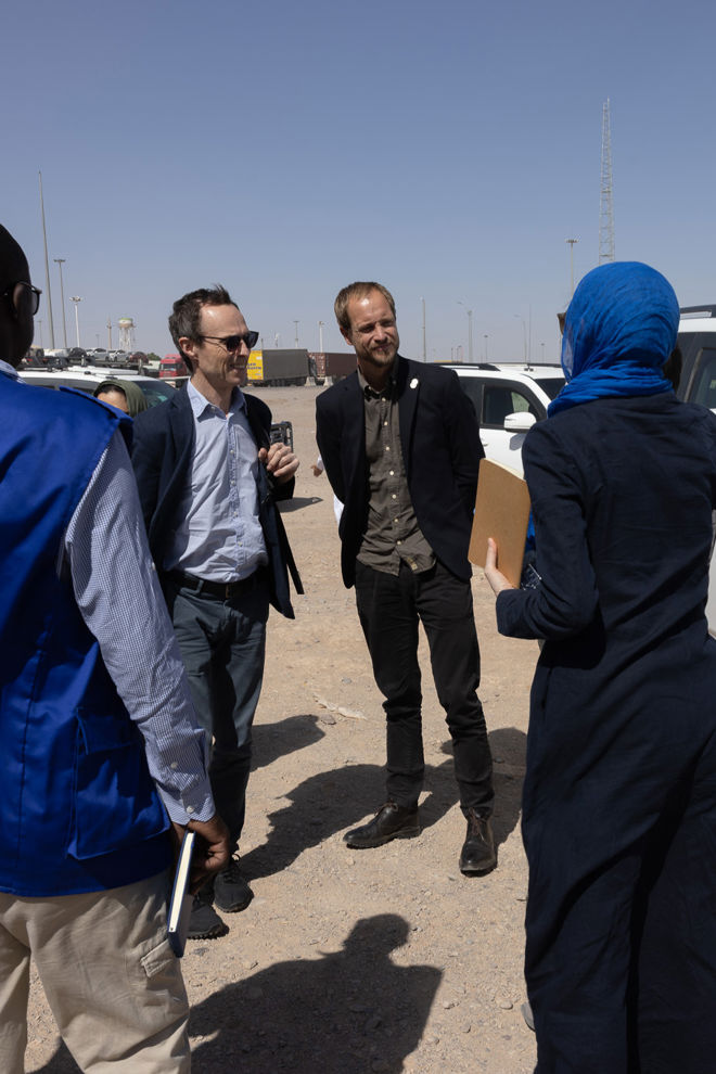 The image shows four people standing in an informal conversation outdoors in a sunny, dusty area. Two of them are men wearing suit jackets and shirts; one is holding a coffee cup and the other has his hands in his pockets. A third person, on the left, is wearing a blue vest and holding a notepad or a document. The fourth person, on the right, is dressed in dark clothing, wears a blue headscarf, and is holding a ring binder or folder.