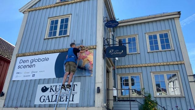 Bildet viser en lys blå, tradisjonell trebygning med hvite vinduskarmer og detaljer i et bymiljø. På veggen henger et stort banner med teksten "Den globale scenen" og logoer fra ulike organisasjoner.