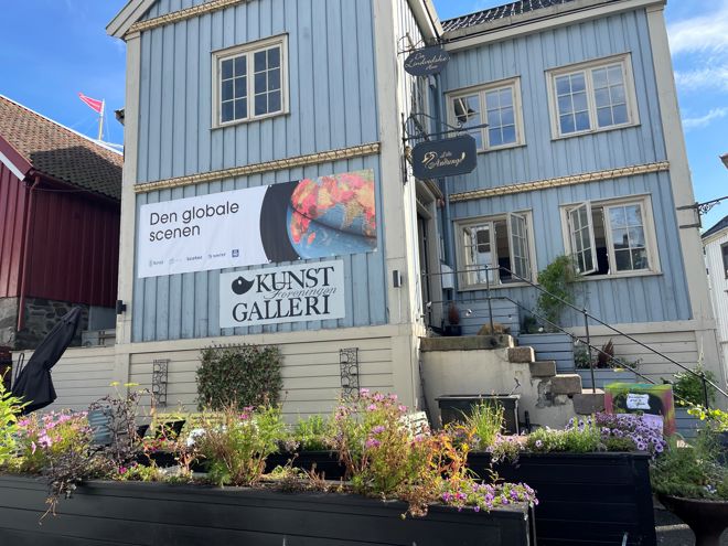 Bildet viser en lys blå, tradisjonell trebygning med hvite vinduskarmer og detaljer i et bymiljø. På veggen henger et stort banner med teksten "Den globale scenen" og logoer fra ulike organisasjoner. 