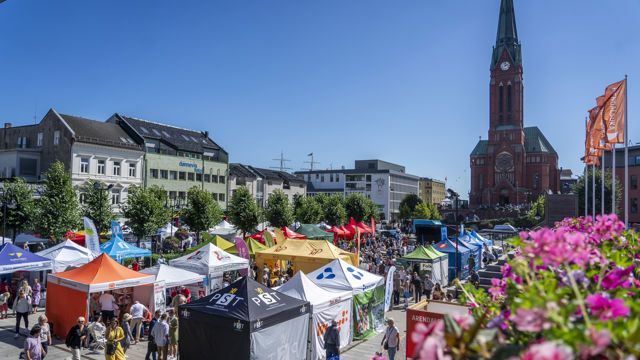 Bildet viser en travel og fargerik scene fra et arrangement utendørs, trolig en festival eller messe, på en solfylt dag. Det er mange forskjellige telt og stands i ulike farger som oransje, gult, hvitt, blått og svart, som representerer ulike organisasjoner og bedrifter. Folk går rundt og besøker teltene, og det er en livlig atmosfære med mange mennesker i alle aldre.
