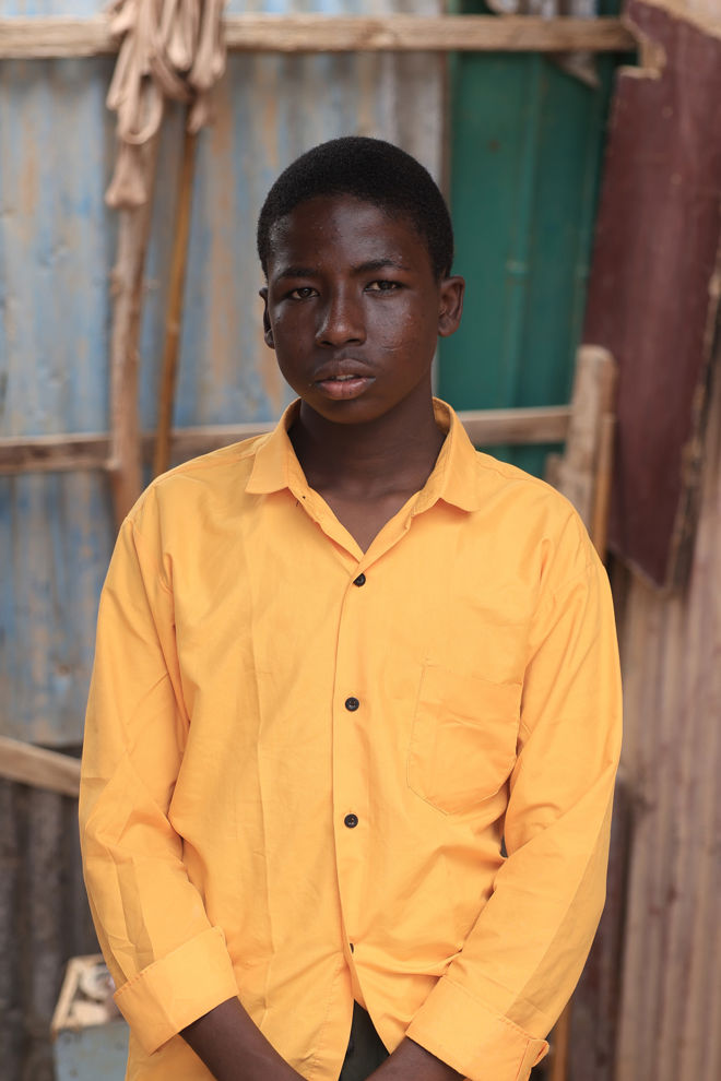 Bildet viser en gutt iført en lys gul skjorte med svarte knapper. Bakgrunnen består av en enkel og slitt utendørs setting, med trelister og metallplater i ulike farger som blått, grønt og brunt.