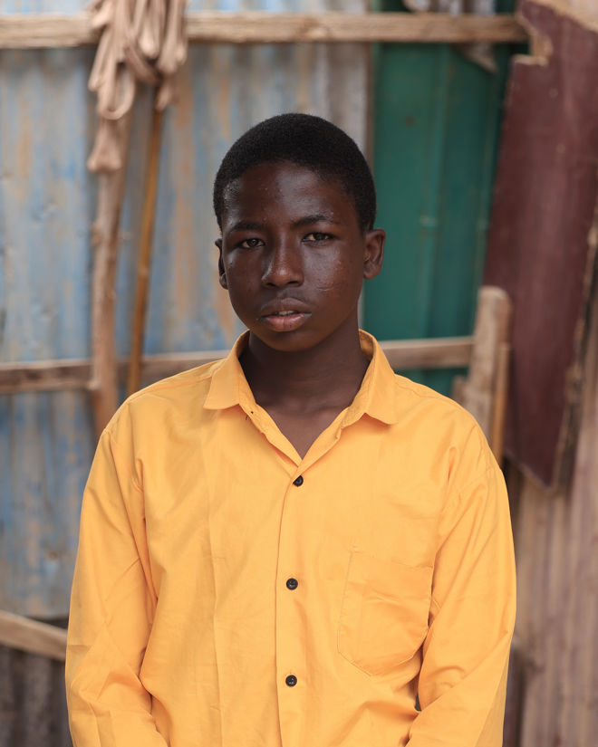 The image shows a boy wearing a bright yellow shirt with black buttons. The shirt has a chest pocket on the left side. The background features a simple outdoor setting with worn wooden planks and corrugated metal panels in shades of blue and brown, possibly part of a shed or similar structure.