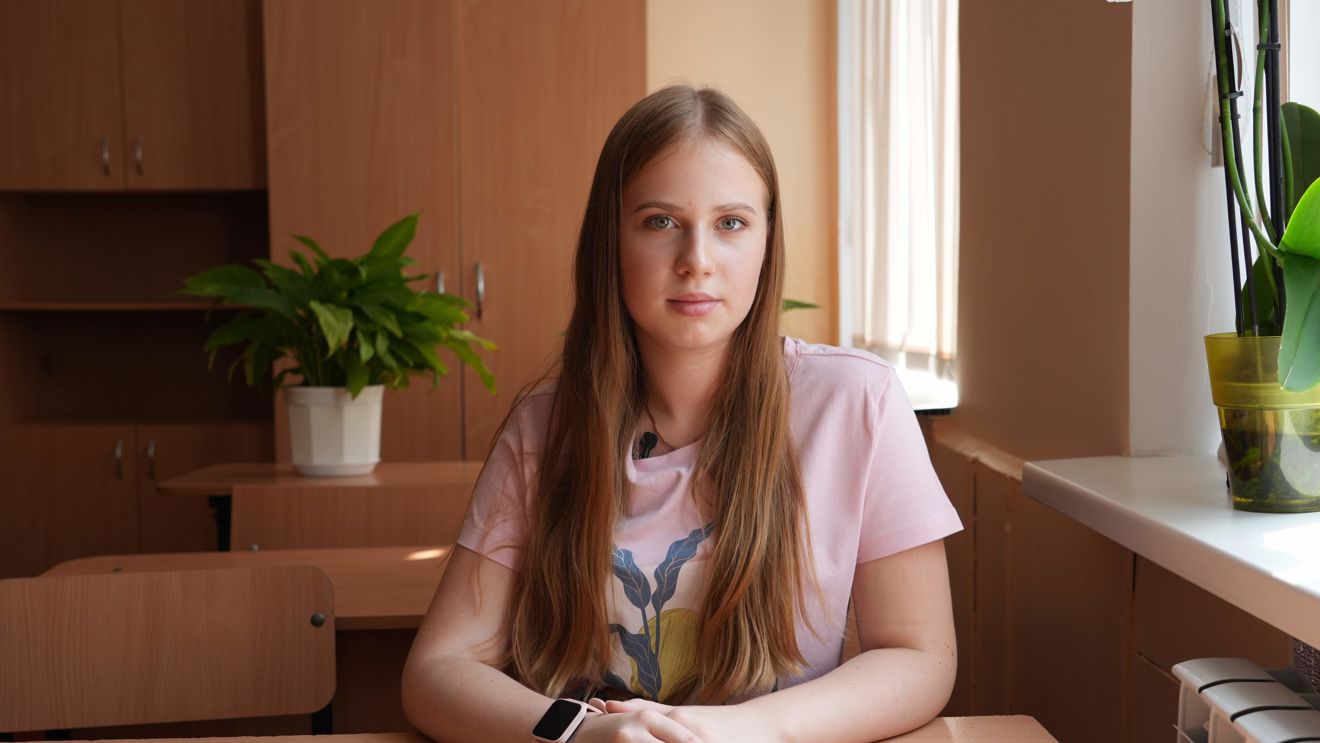 The image shows a teenage girl seated at a wooden desk in a bright room. They are wearing a light pink t-shirt with a graphic design.