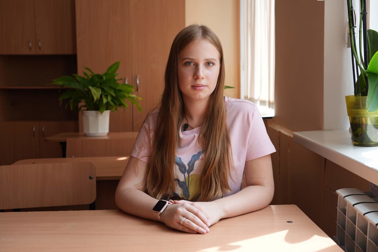The image shows a teenage girl seated at a wooden desk in a bright room. They are wearing a light pink t-shirt with a graphic design.