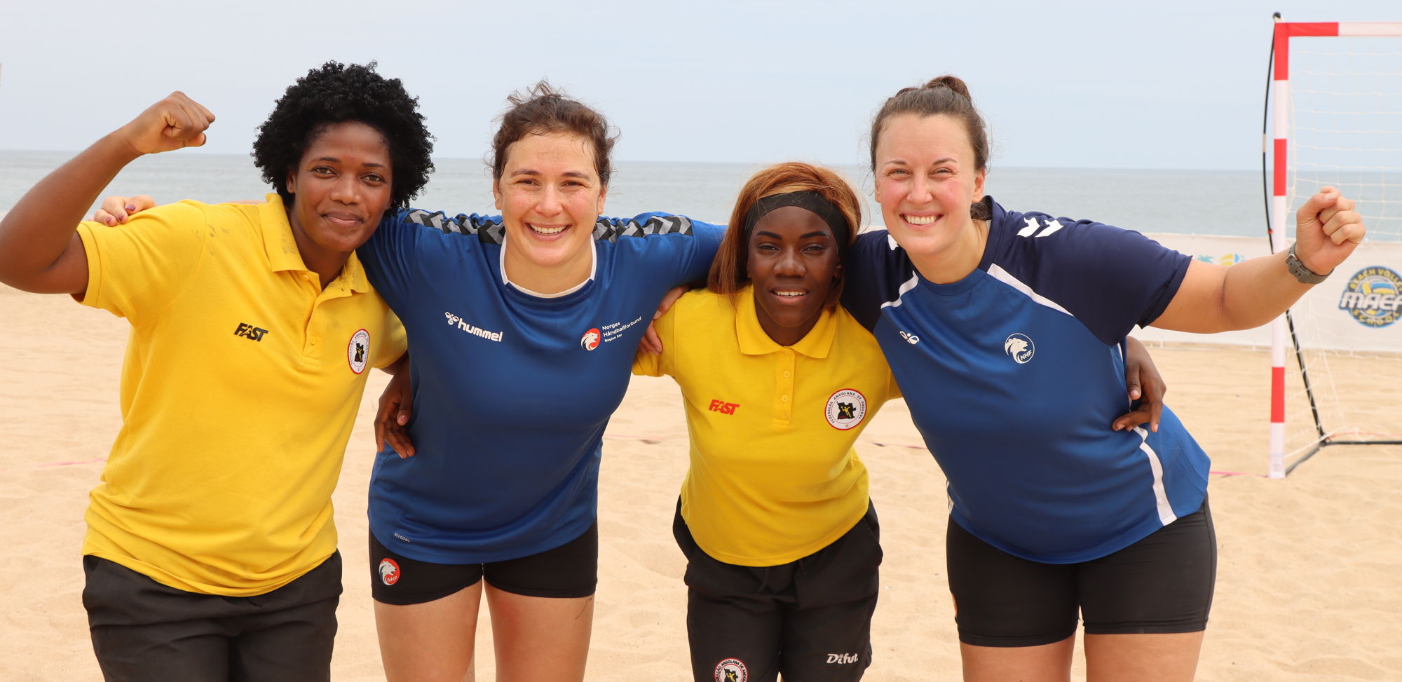 Four woman are standing close together on a sandy beach, with their arms around each other's shoulders in a friendly pose. Two are wearing yellow sports shirts and black shorts, while the other two are in blue sports shirts and black shorts. A goalpost can be seen in the background, and the ocean is visible behind them under a cloudy sky.