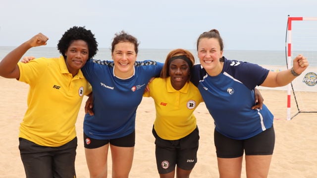 Fire personer står tett sammen på en sandstrand foran et håndballmål, med armene rundt hverandre og smilende kroppsspråk. To av dem har på seg gule trøyer med logo, mens de to andre har blå trøyer – også med ulike logoer. De holder armene opp i en energisk og seiersbevis gest, og bak dem kan man se havet og en lys, overskyet himmel. Bildet utstråler samhold, glede og sportslig lagånd.
