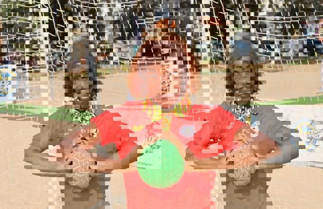 A woman stands on a sandy sports field in front of a goal net, wearing a bright red sports shirt with a colorful patterned collar. They are holding a green ball with both hands, positioned at chest height. The background shows some trees, parked cars, and banners, indicating an outdoor event or tournament setting. The overall atmosphere is sporty and energetic.