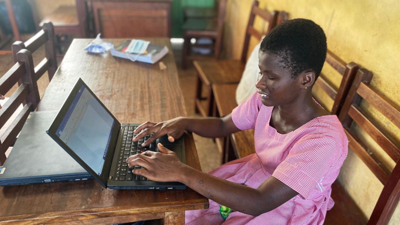 The image shows a girl sitting at a wooden table working on a laptop. The person is wearing a pink and white striped dress. On the table, there is also another closed laptop and some books or notebooks.