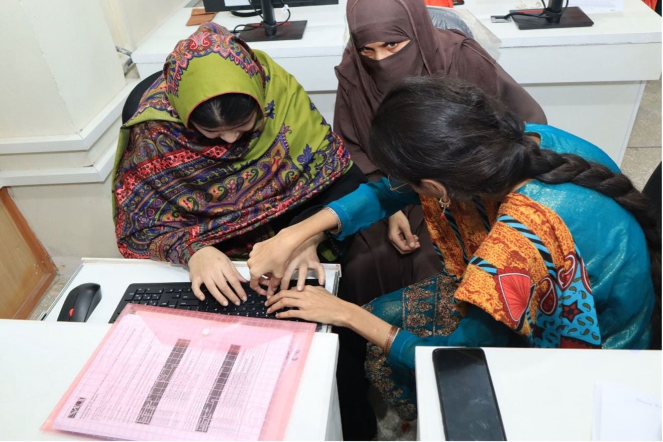 The image shows three women collaborating at a table with a computer. Two of them are pointing and working together on the keyboard. One is wearing a colorful green shawl with patterns, while the other is dressed in a blue and orange outfit with a patterned design. On the table, there is also a pink folder with a document inside and a mobile phone.