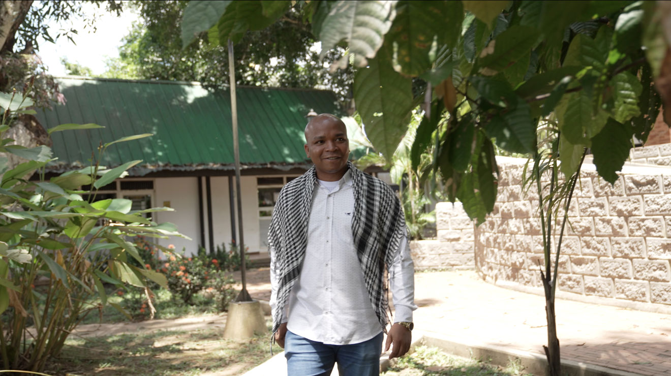A man is walking outside. Behind him, we see a small white house, and in front of him stands a green tree.