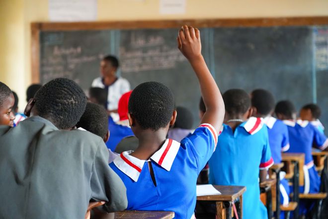 The image shows a classroom with children sitting at desks, seen from behind. The children are wearing blue school uniforms with white and red details. One student in the foreground has their hand raised, clearly engaged in the lesson. At the front of the room, a teacher stands by a large blackboard with writing on it. The atmosphere is focused, and the room is filled with students paying attention to what is happening.