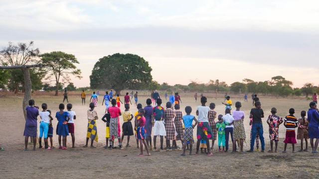 Bildet viser en stor gruppe barn og ungdommer som står ute på en åpen, støvete slette i Zambia. De har ryggen til kameraet og ser på en fotballkamp eller lek som foregår lenger fremme. Barna har på seg fargerike klær, og stemningen er livlig og sosial. Rundt sletta vokser det noen spredte trær, og lyset er mykt, som ved solnedgang eller tidlig morgen.
