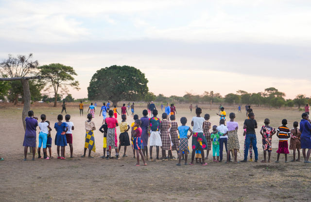 The image shows a large group of children and young people standing outside on an open, dusty field in Zambia. They have their backs to the camera and are watching a football match or game taking place further ahead. The children are dressed in colorful clothes, and the atmosphere is lively and social. There are a few scattered trees around the field, and the light is soft, as if it is sunset or early morning.