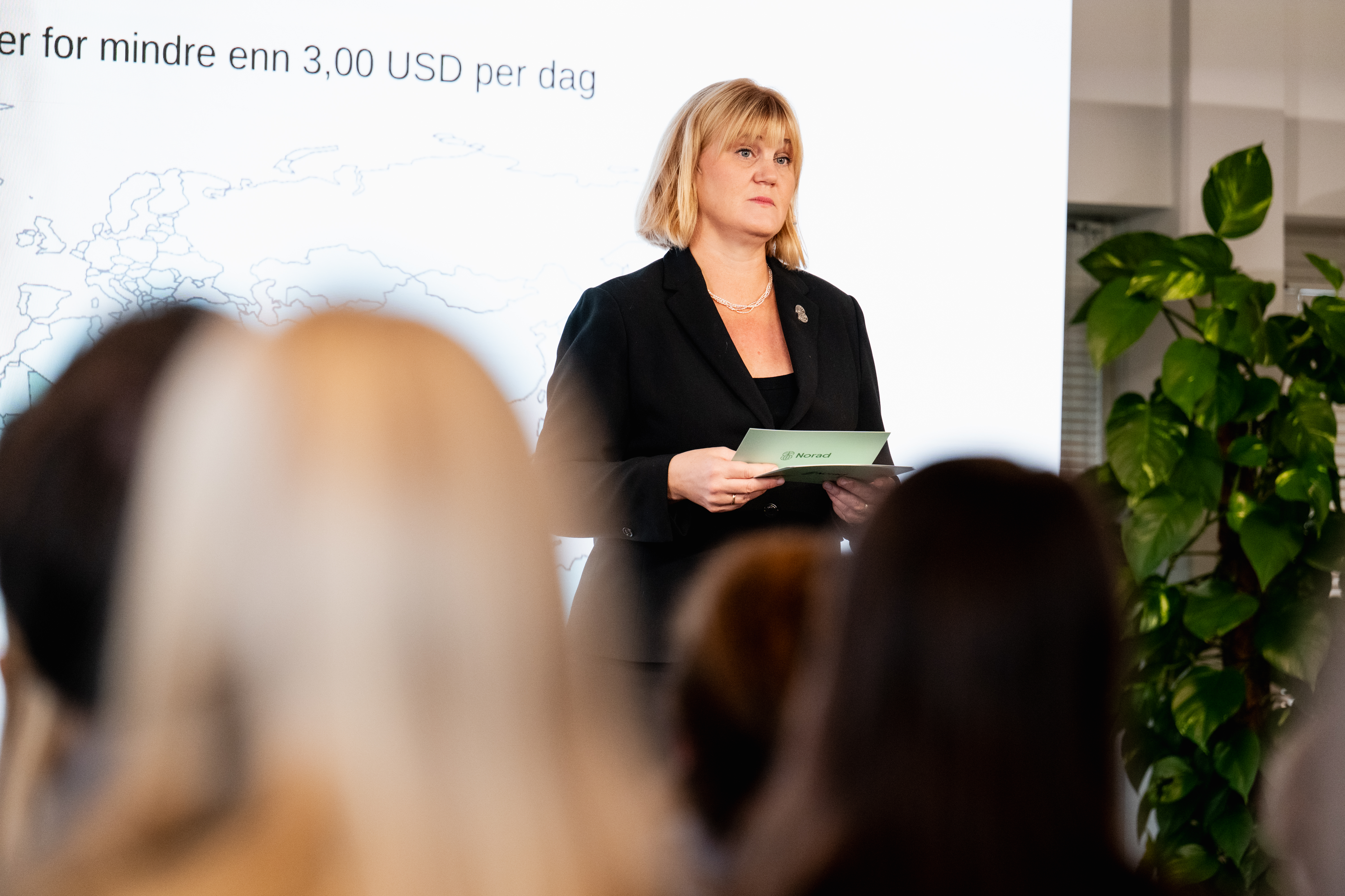 Bildet viser en kvinne som står foran en forsamling innendørs,  under en presentasjon. Hun har skulderlangt lyst hår og er kledd i en mørk dressjakke med en enkel topp under, samt et halskjede.