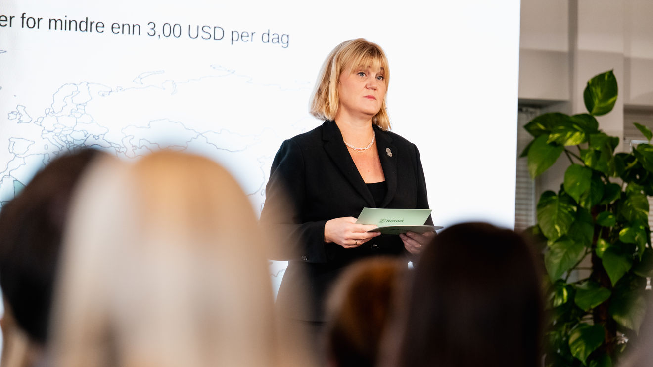 Bildet viser en kvinne som står foran en forsamling innendørs, under en presentasjon. Hun har skulderlangt lyst hår og er kledd i en mørk dressjakke med en enkel topp under, samt et halskjede.