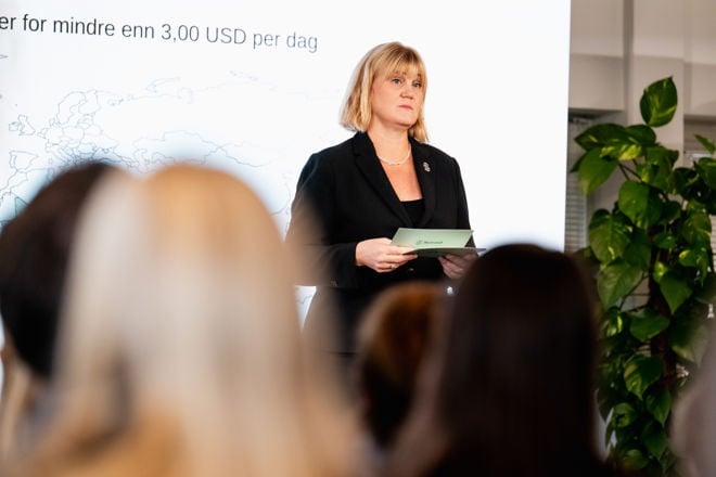 Bildet viser en kvinne som står foran en forsamling innendørs, under en presentasjon. Hun har skulderlangt lyst hår og er kledd i en mørk dressjakke med en enkel topp under, samt et halskjede.