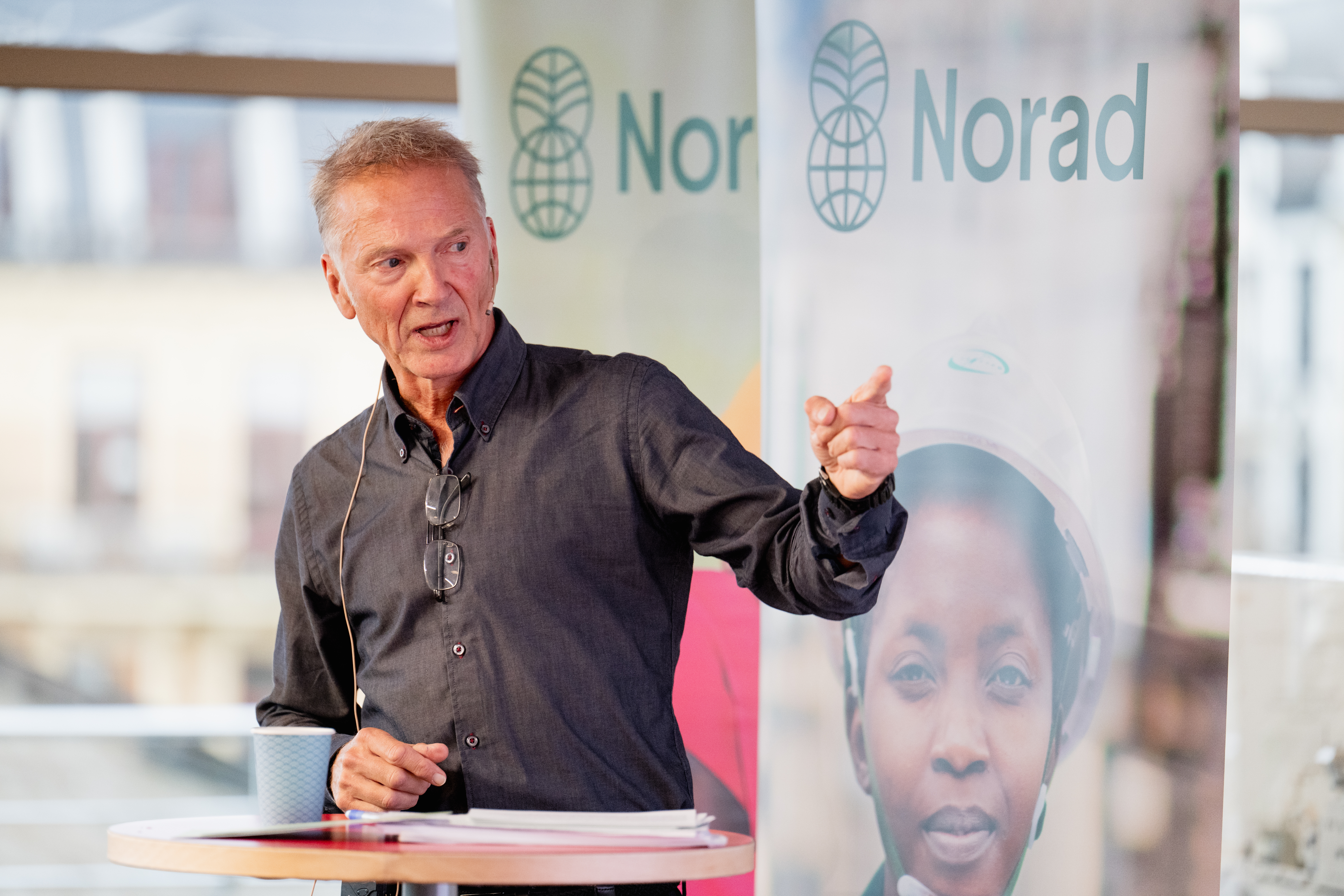 En mann står ved et rundt bord og holder en presentasjon. Han har på seg mørk skjorte, briller henger rundt halsen, og han bruker mikrofon. På bordet foran ham ligger papirer og en kopp. Bak ham er det Norad-bannere og store vinduer med utsikt til byen.
