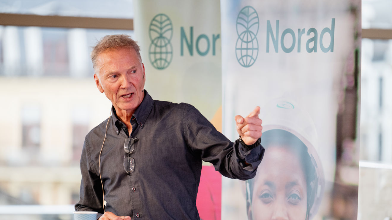 En mann står ved et rundt bord og holder en presentasjon. Han har på seg mørk skjorte, briller henger rundt halsen, og han bruker mikrofon. På bordet foran ham ligger papirer og en kopp. Bak ham er det Norad-bannere og store vinduer med utsikt til byen.