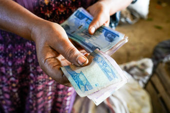 The image shows a person holding and counting banknotes with both hands. The person has dark skin and is wearing a dress with a purple and pink patterned design. The banknotes are of different denominations, and it appears they are being used for a payment or transaction. 