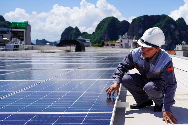 En person med hjelm og arbeidsklær inspiserer solcellepaneler på et tak med fjell og bygninger i bakgrunnen.