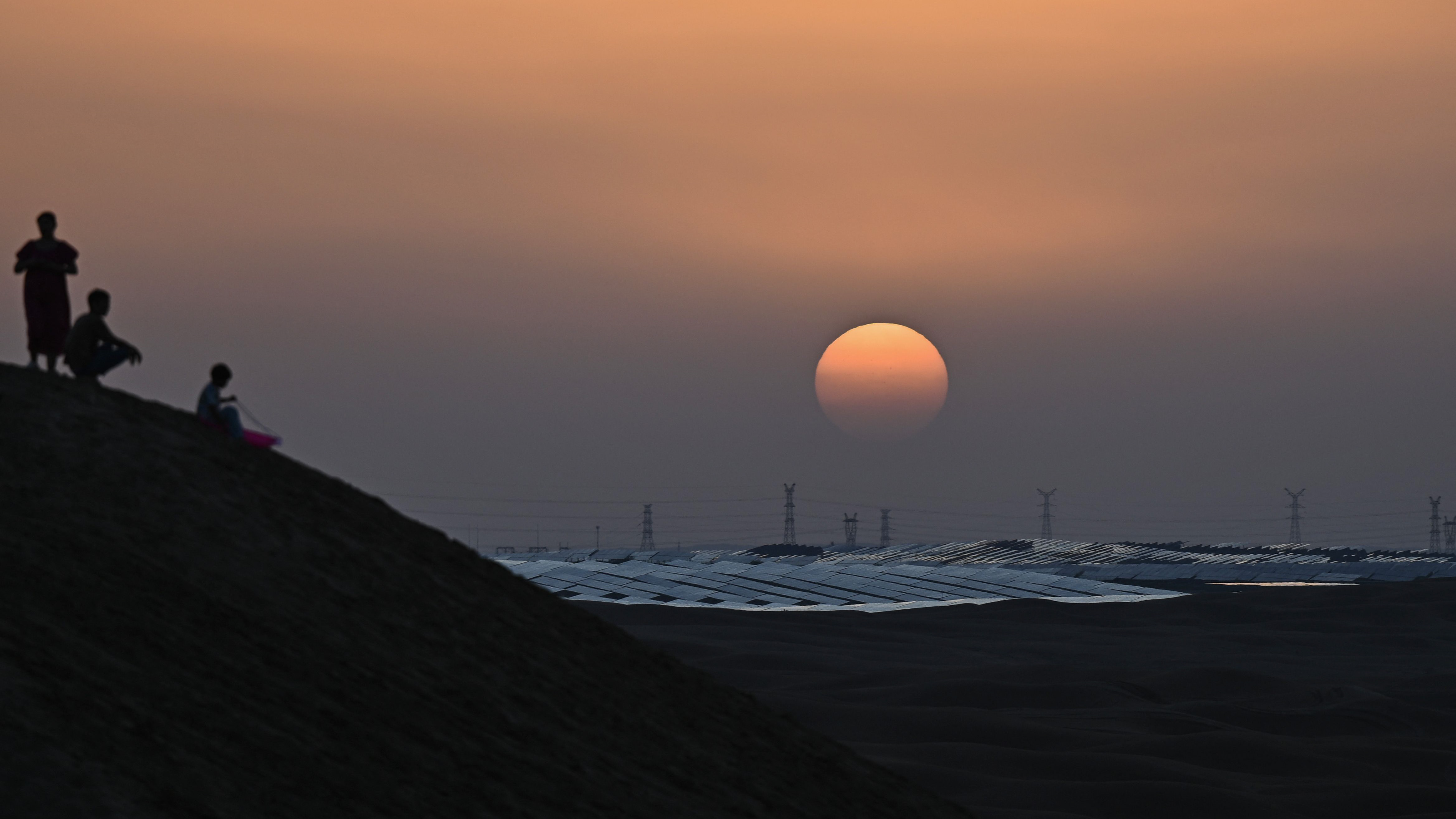 Silhuetter av tre personer står på en sanddyne i solnedgang med et stort solcelleanlegg i bakgrunnen.
