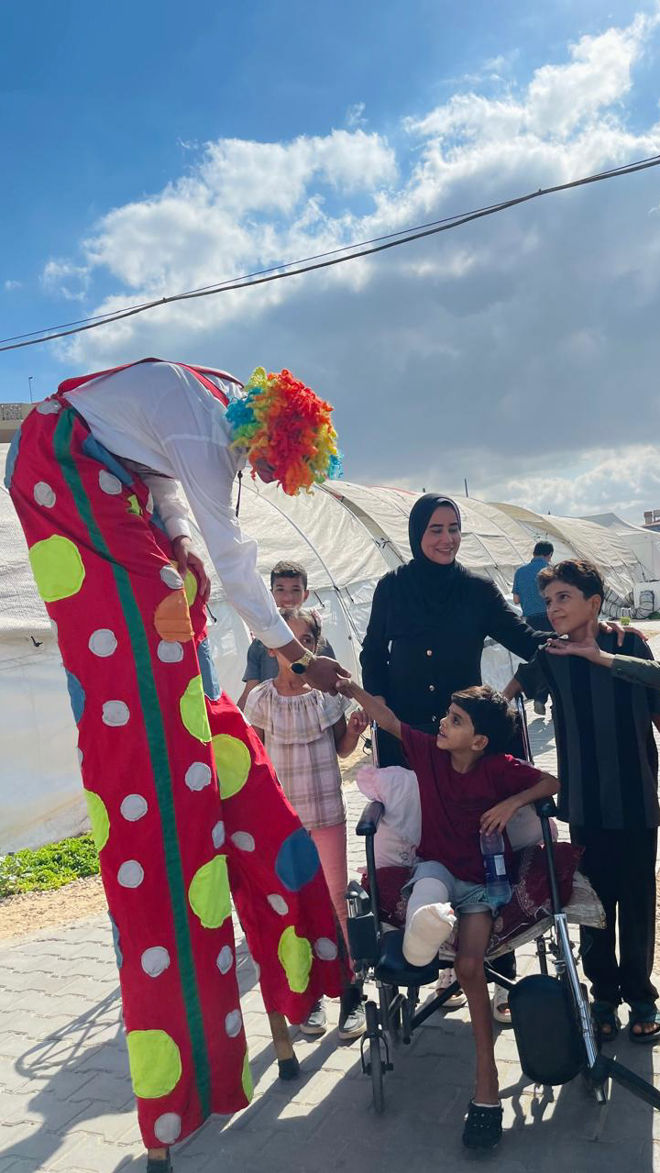 A person dressed as a clown on stilts interacts with children, one of whom is sitting in a wheelchair, outdoors near tents.