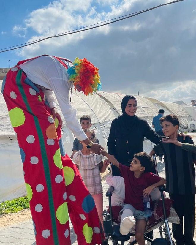A person dressed as a clown on stilts interacts with children, one of whom is sitting in a wheelchair, outdoors near tents.