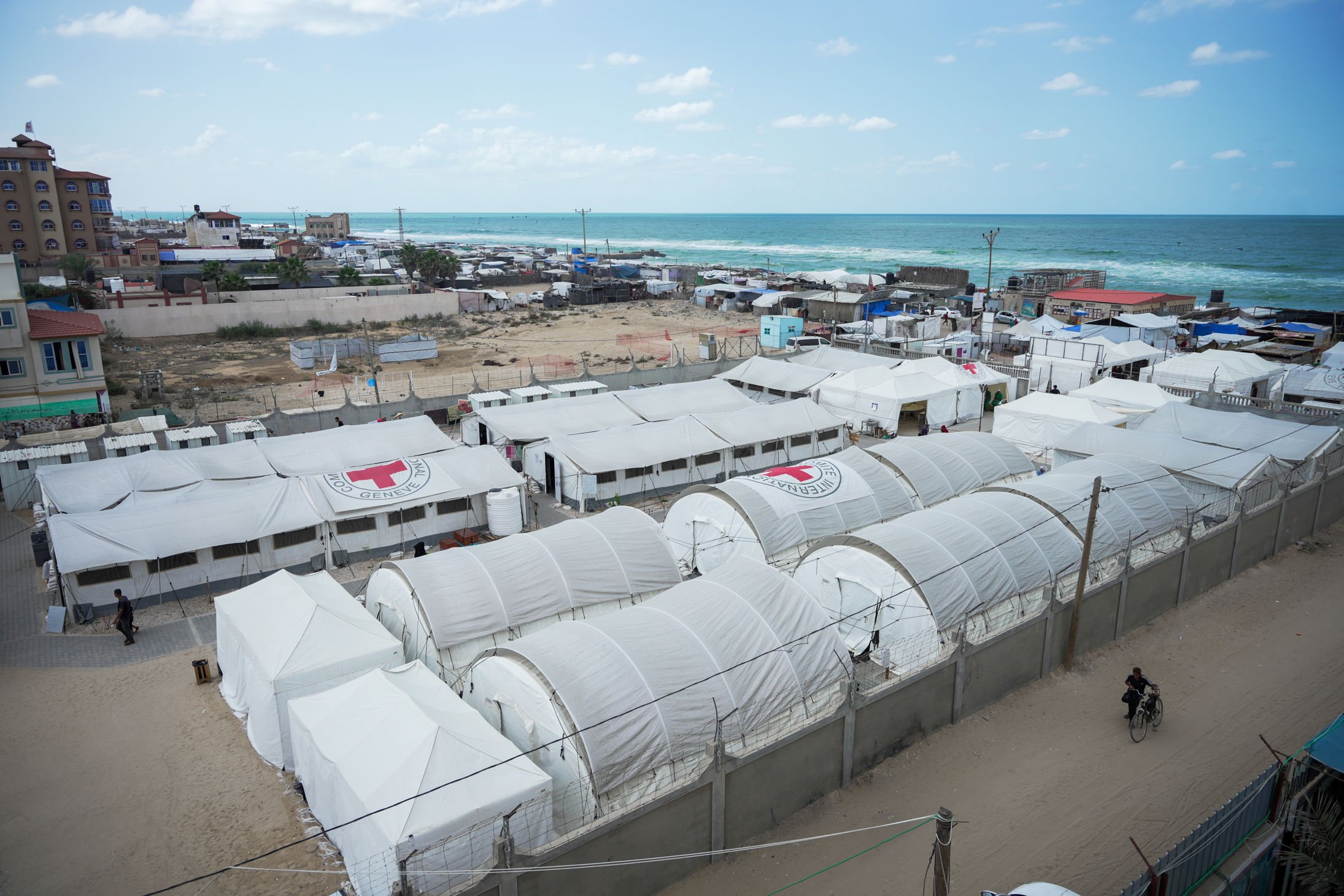 A large field hospital with white tents and Red Cross symbols is set up near the coastline, with a few people walking and buildings in the background.