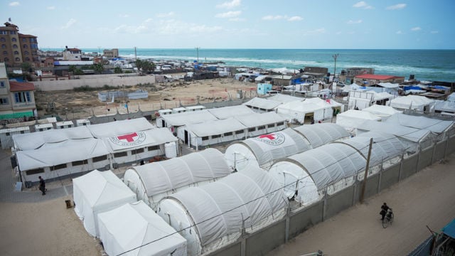 Et stort feltsykehus med hvite telt og Røde Kors-symboler er satt opp nær kysten, med noen personer som går og bygninger i bakgrunnen.