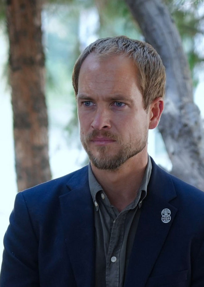 Portrett av Erik Abild. Han står med kort lyst hår iført skjorte og mørk jakke med Norad-logo på en nål, utendørs foran trær.