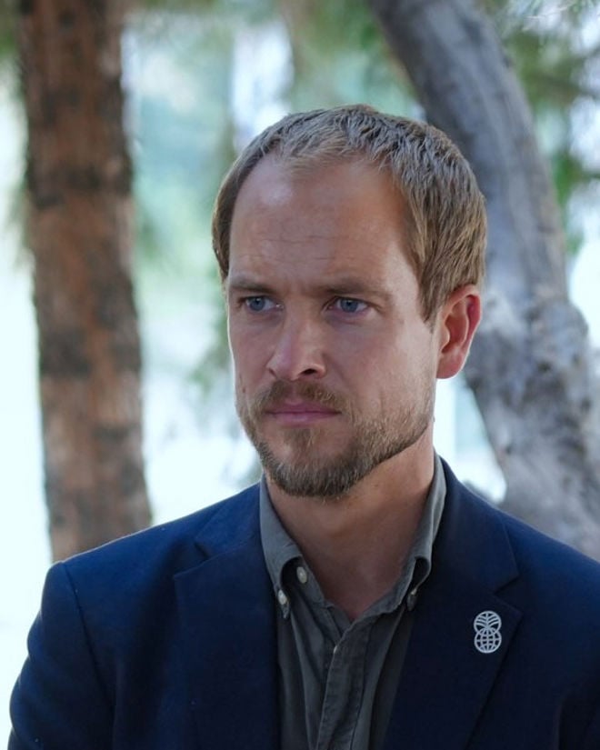 Portrait of Erik Abild. He stands with short blond hair wearing a shirt and dark jacket with a Norad logo pin, outdoors in front of trees.