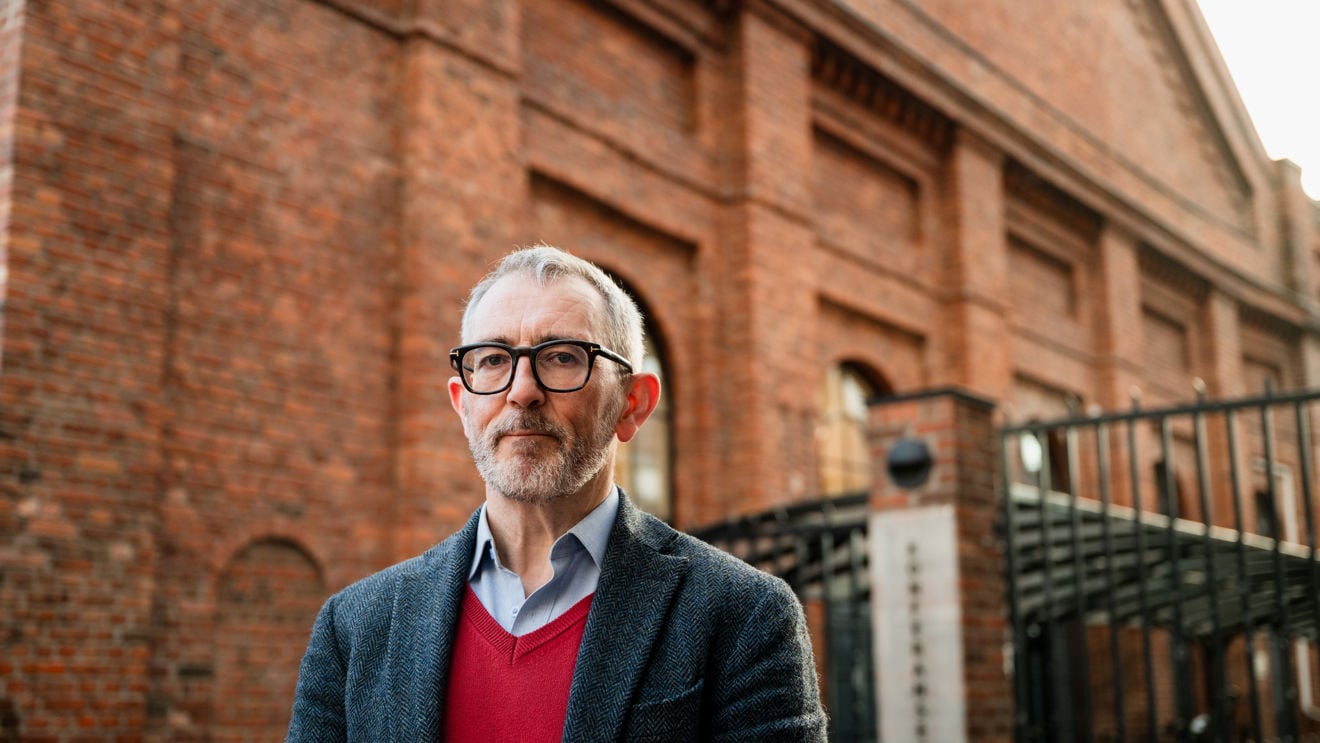 A man with grey hair and glasses, wearing a red sweater and dark blazer, stands outside in front of a large brick building