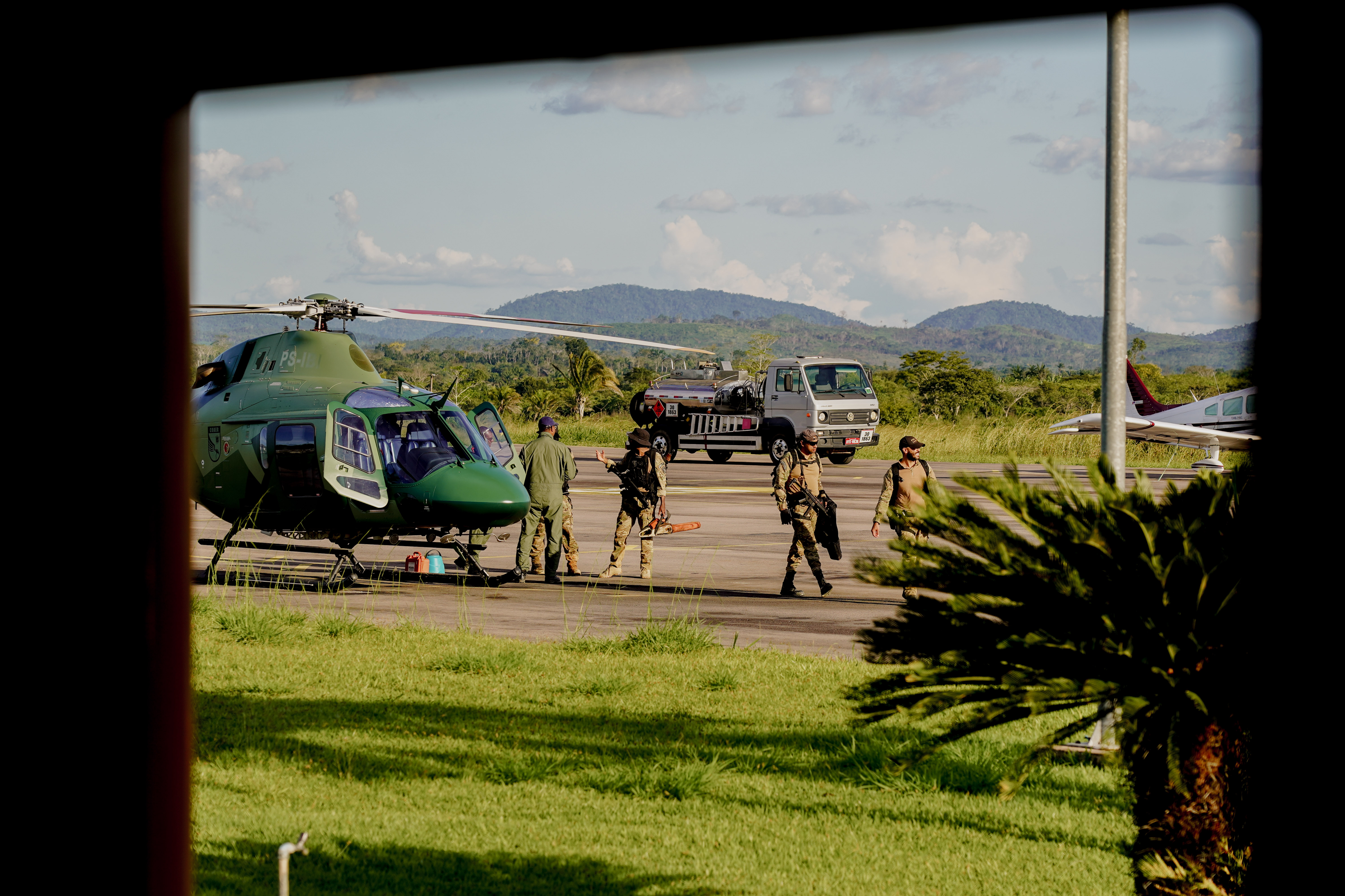 The image shows a group of people in military or police uniforms at an airport. They are standing next to a green helicopter, and there is also a car and a small aircraft in the background. The surrounding landscape features green grass, palm trees, and distant mountains beneath a blue sky with a few clouds