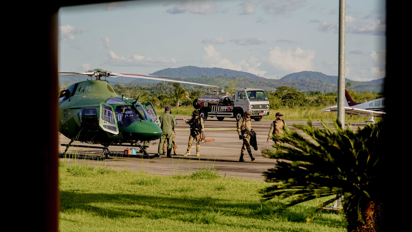 The image shows a group of people in military or police uniforms at an airport. They are standing next to a green helicopter, and there is also a car and a small aircraft in the background. The surrounding landscape features green grass, palm trees, and distant mountains beneath a blue sky with a few clouds