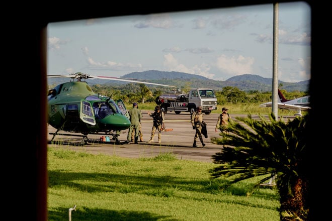 Bildet viser en gruppe personer i militær- eller politiuniformer på en flyplass. De står ved siden av et grønt helikopter og det er også en bil og et småfly i bakgrunnen. Landskapet rundt består av grønt gress, palmer og fjell i det fjerne under en blå himmel med noen skyer.
