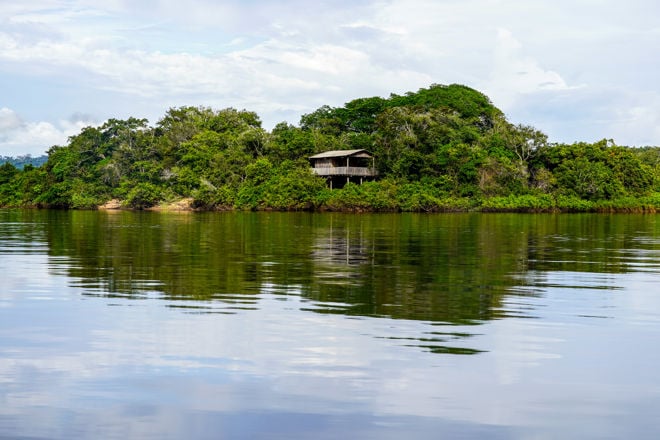 Bildet viser en frodig, grønn øy med tett vegetasjon. Midt på øya står et enkelt, trekledd hus på påler, delvis skjult av trærne. Øya er omgitt av stille vann som speiler både hus og vegetasjon. Himmelet er lys med noen skyer, og landskapet gir en følelse av ro og isolasjon, langt fra by eller tettsted.