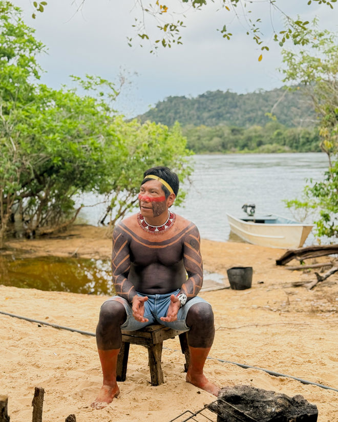 A man is sitting on a simple wooden chair on a sandy riverbank. His upper body, arms, and legs are covered in extensive black and red body paint patterns.