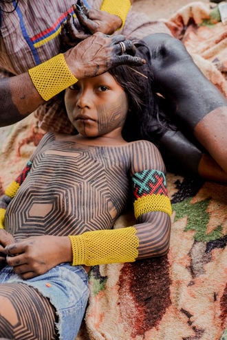 Bildet viser et barn som ligger på et teppe, mens en voksen kvinne sitter ved siden av og steller barnets hår. Barnet har geometriske, sorte kroppsmalinger over hele overkroppen, armene og bena, og har på seg dongerishorts.