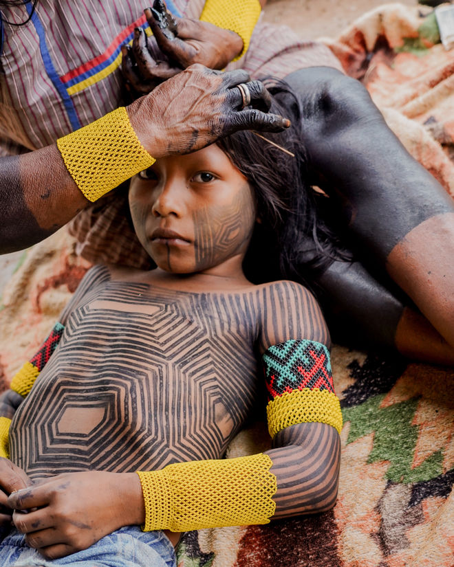 The image shows a child lying on a blanket, while an adult woman sits beside them, tending to the child's hair. The child has geometric black body paintings covering the torso, arms, and legs, and is wearing denim shorts.
