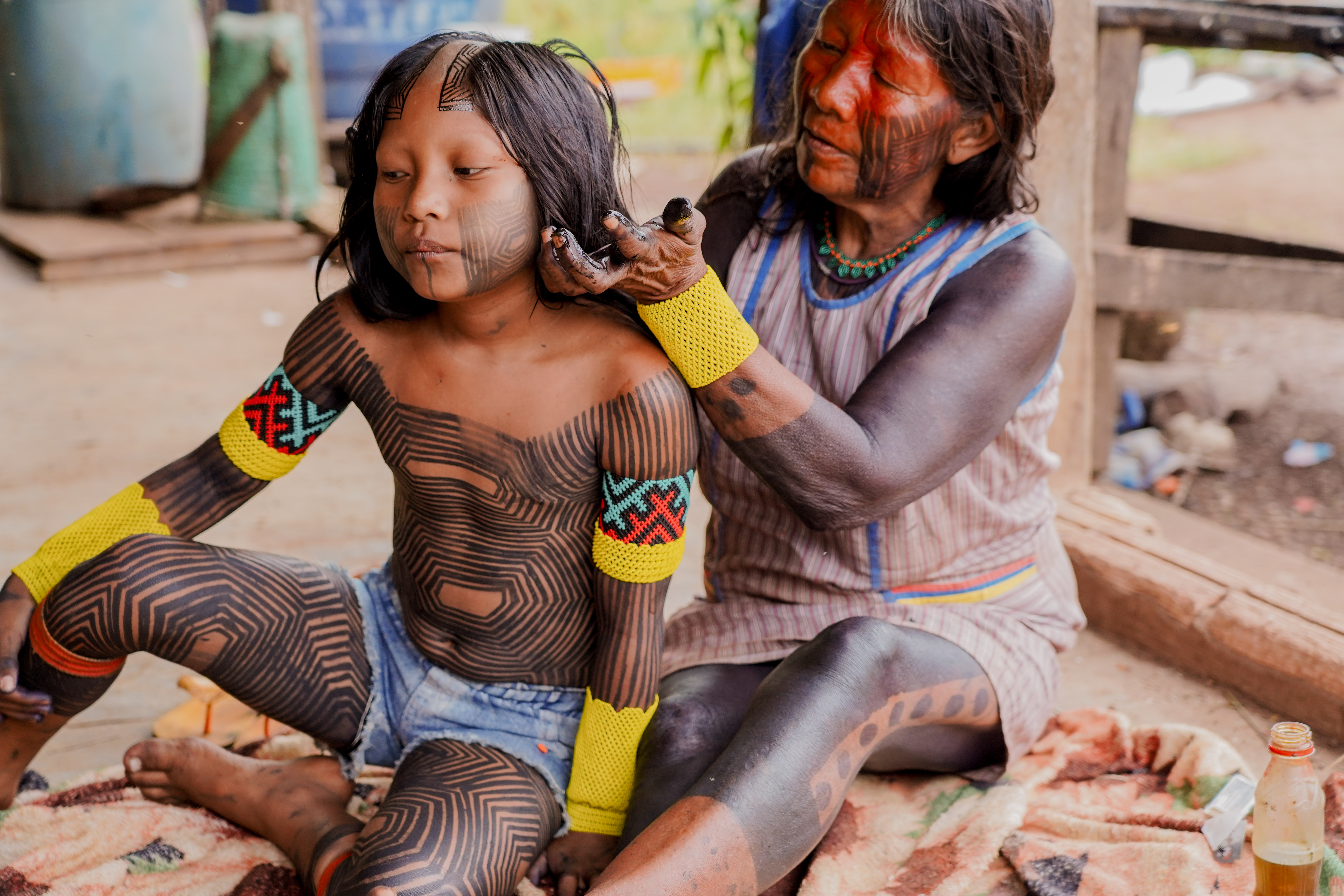 The image shows two people, an adult woman and a child, sitting on a blanket outdoors. The woman is holding and tending to the child's hair. Both have their bodies adorned with extensive black patterns that resemble traditional body painting.
