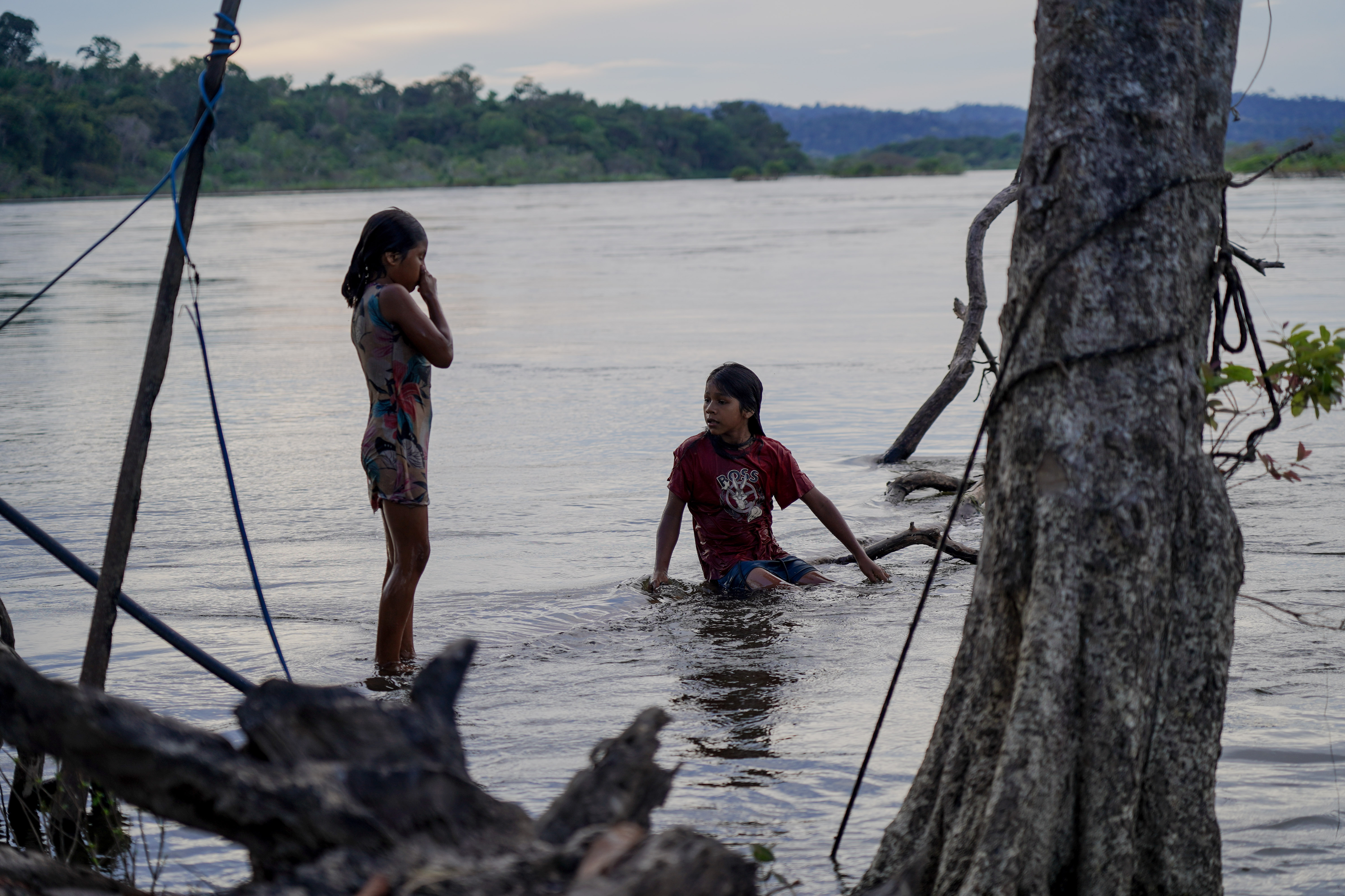 The image shows two children by the riverbank in a natural landscape. One is standing in the water wearing a short summer dress, while the other is squatting in the water wearing a red T-shirt and shorts. The water is calm, and there are trees and vegetation around them, with a forest in the background