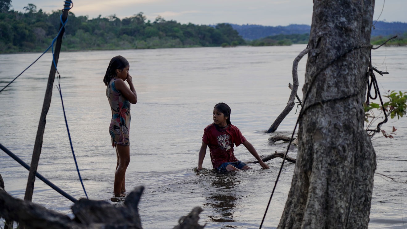 The image shows two children by the riverbank in a natural landscape. One is standing in the water wearing a short summer dress, while the other is squatting in the water wearing a red T-shirt and shorts. The water is calm, and there are trees and vegetation around them, with a forest in the background