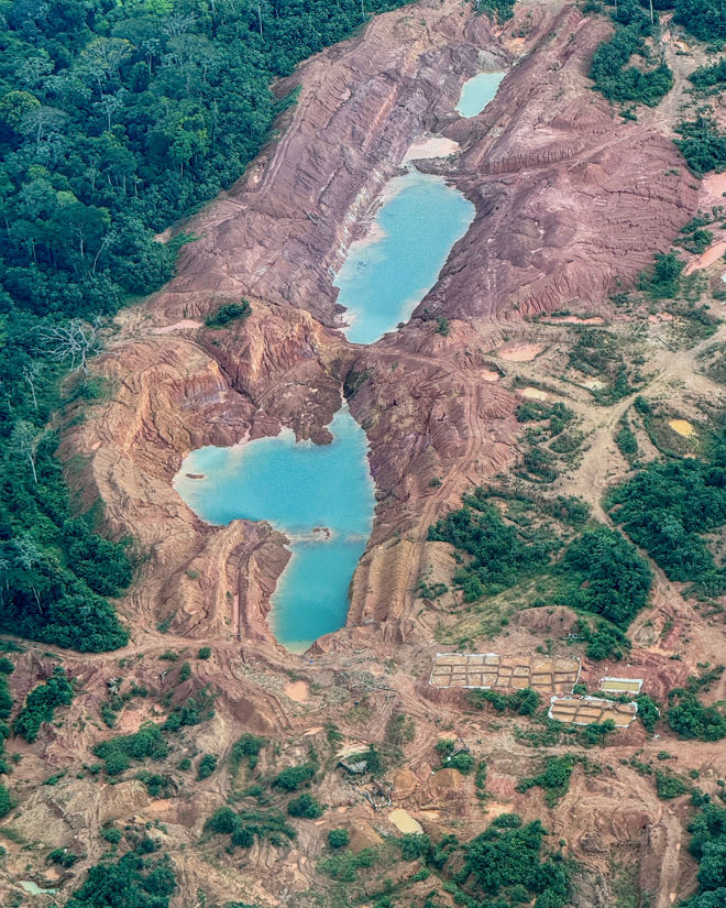 The image shows a large area of deforested and excavated land, likely an open-pit or gold mine in the rainforest. In the middle of the site are several large blue-green ponds surrounded by red and brown soil, with remnants of forest along the edges