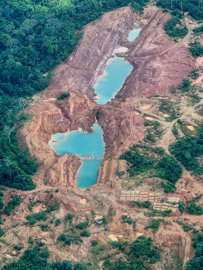 Bildet viser et stort område med avskoget og oppgravd landskap, sannsynligvis et dagbrudd eller en gullgruve i regnskogen. Midt i området ligger flere store, blågrønne dammer omgitt av rødt og brunt jord, og det er rester av skog rundt kanten.