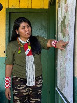 En kvinne står foran en vegg og peker på et stort kart som henger opp. Hun har langt, mørkt hår og bærer en grønn T-skjorte med et brasiliansk flagg på ermet, kamuflasjemønstrede bukser, og fargerike armbånd i rødt og hvitt.